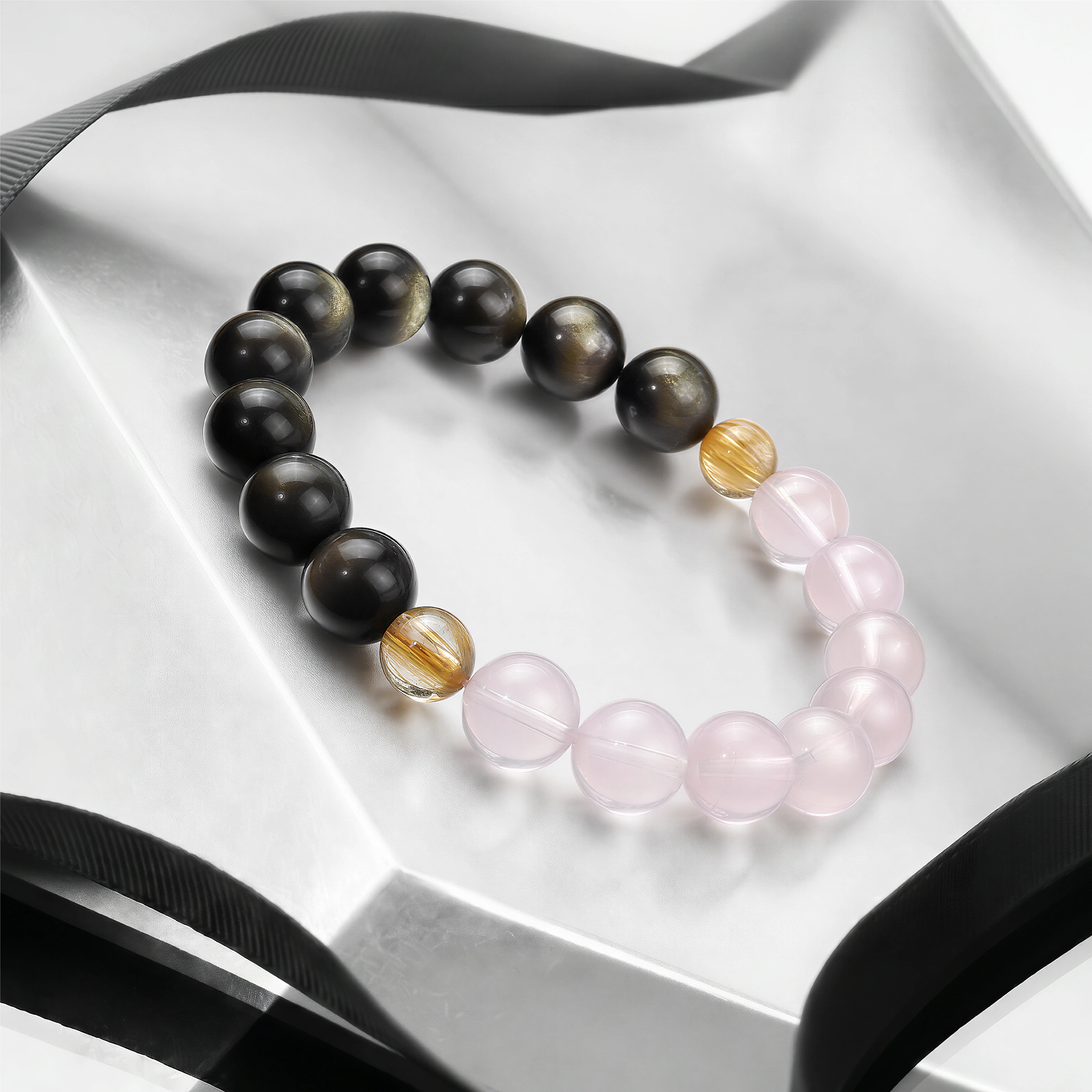 The Obsidian, Pink Quartz and Rutilated Quartz Bracelet on a reflective white surface, BE.