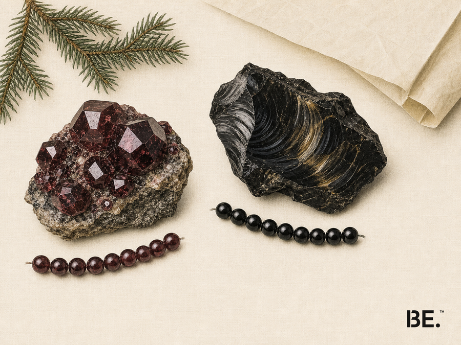 Editorial illustration on warm ivory paper showing a deep-red crystal cluster, a black glassy stone with gold-coloured sheen, short rows of burgundy and black beads, evergreen foliage and the BE. logo