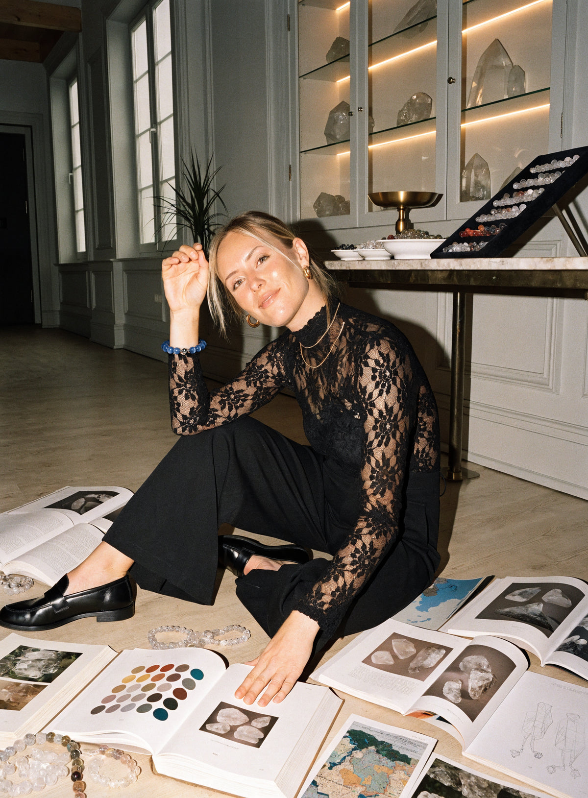 Woman in a black lace top seated among open mineral reference books and bead strands.