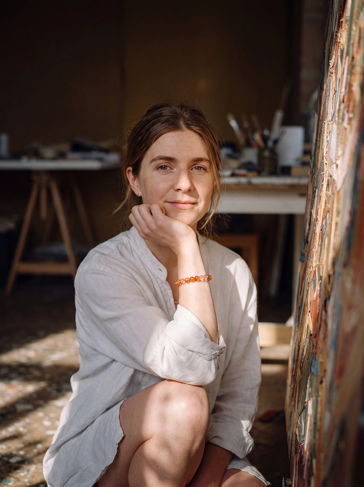 Artist seated beside a painted canvas, wearing an amber bead bracelet.