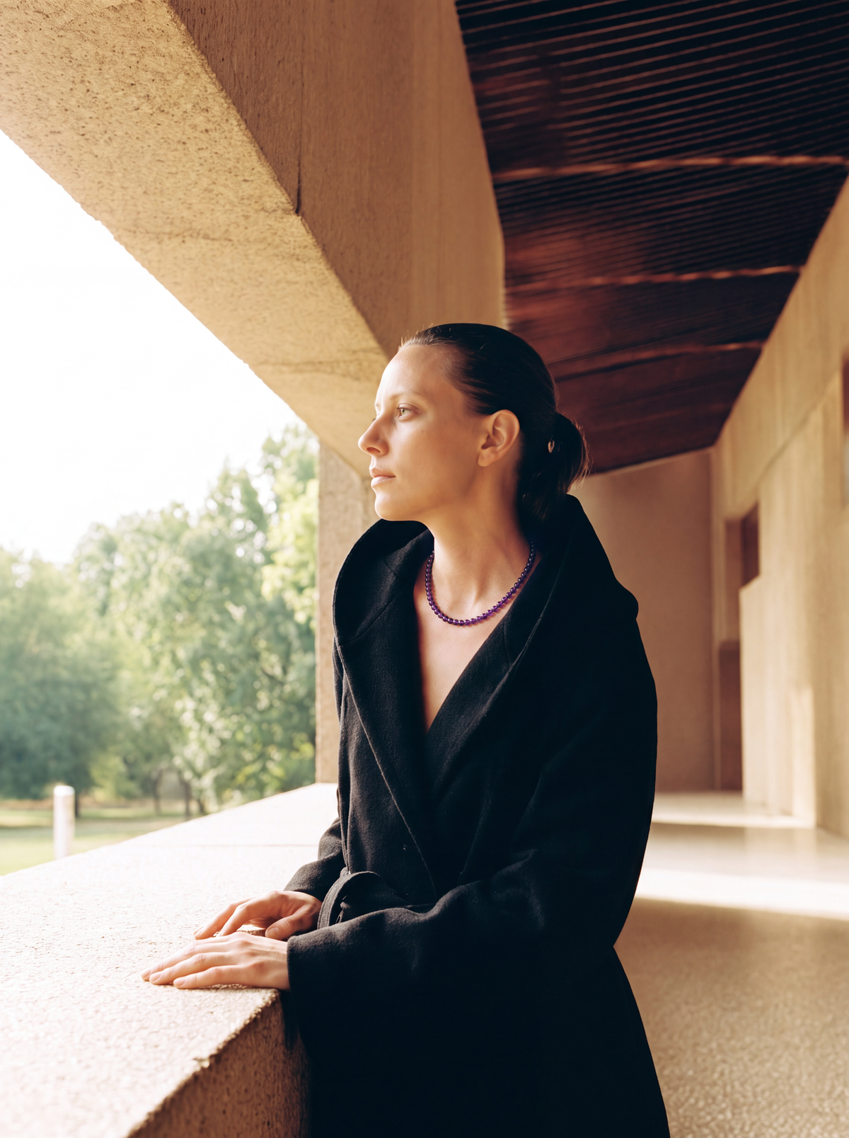 Woman in a black coat wearing a purple bead strand beside a concrete window.