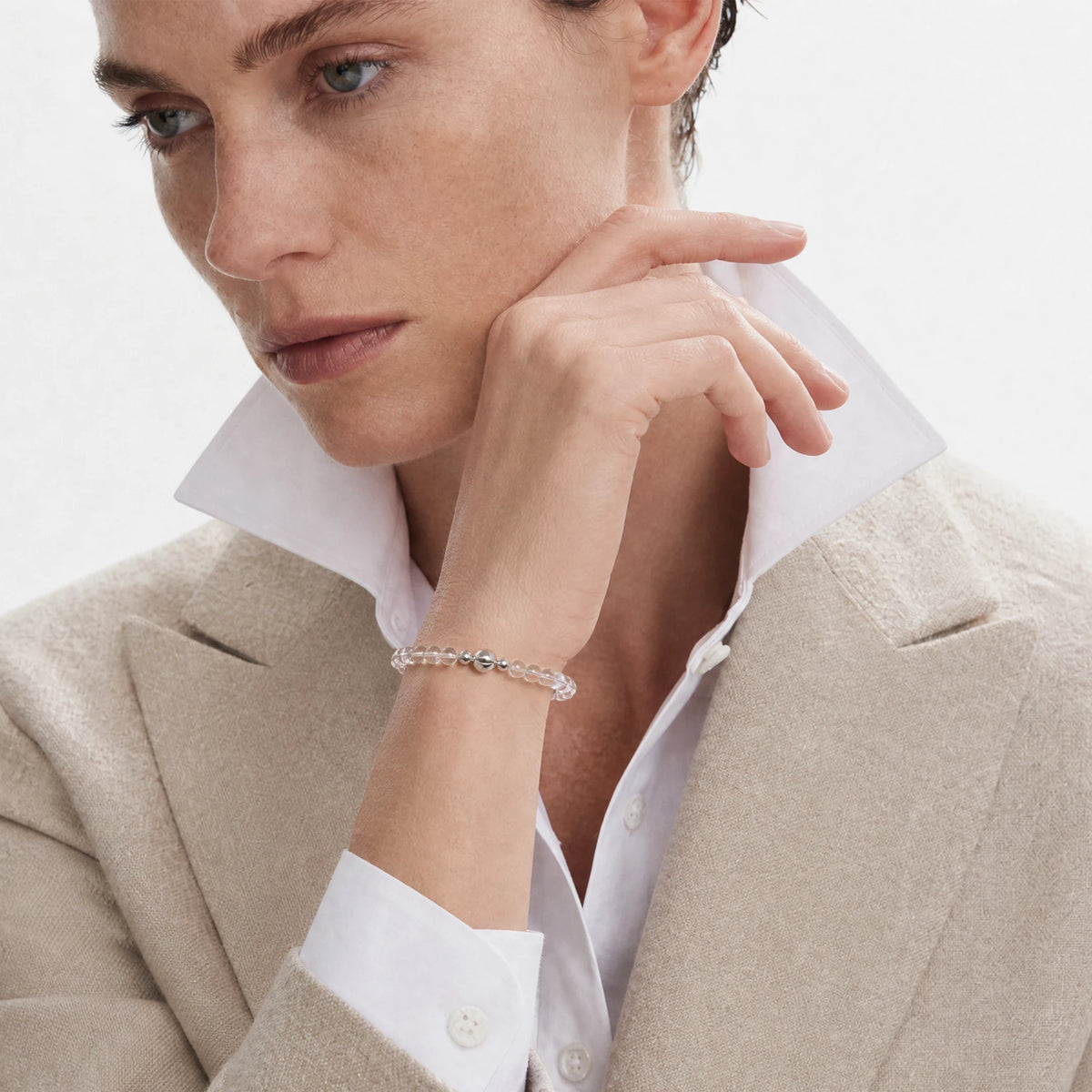 Clear Quartz 6mm Strand Bracelet worn on the wrist with a beige jacket and white shirt, hand raised near the face.