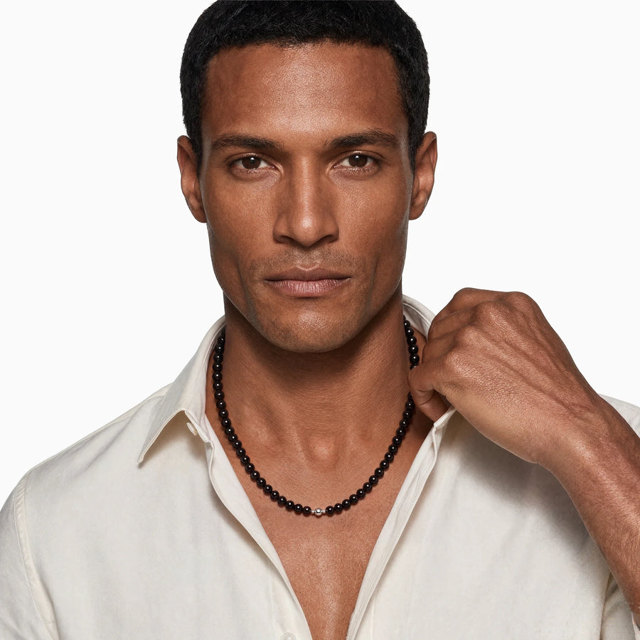 Black onyx bead necklace worn by a man in an open white shirt, with one hand near his collar.