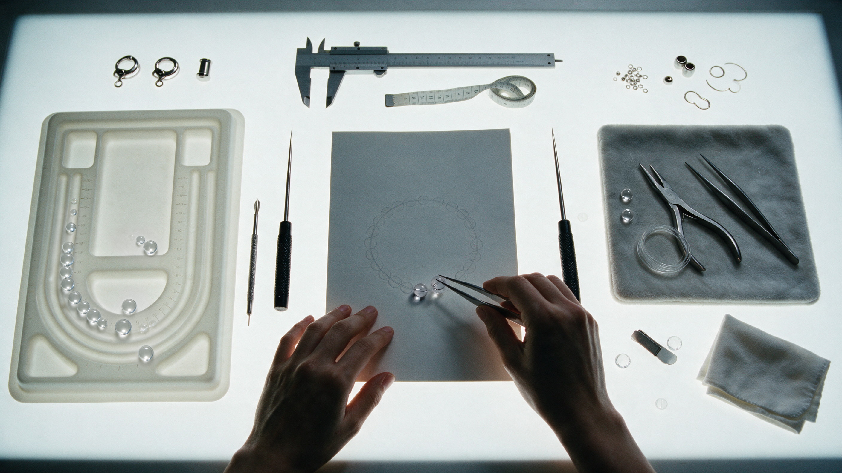 Hands above an open record book, surrounded by bead trays and jewelry tools.